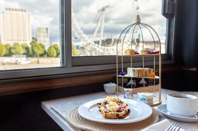 A scone with cream and a tiered tea stand on a table by a boat window, the London Eye outside